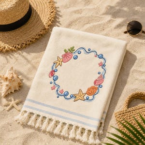 May include: A cream-colored beach towel featuring an embroidered circular wreath design of pink and orange seashells, yellow starfish, and blue bubbles. The towel has two horizontal light blue stripes near the bottom edge and white fringe trim.