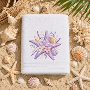 Peut inclure: Une serviette rectangulaire blanche ornée d'une broderie représentant une étoile de mer violette décorée de petites fleurs, de brins de lavande et de coquillages. La serviette présente une texture avec des bandes horizontales en bas.