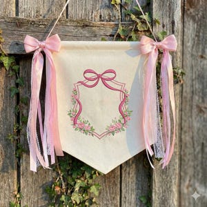 May include: A cream banner with a pink bow and ribbon design. The banner features a pink ribbon frame with a floral wreath and is adorned with pink bows and ribbons for hanging. The banner is hanging on a wooden surface.