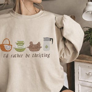 Puede incluir: Sudadera beige con ilustraciones bordadas de una cesta, cuencos, una gallina y una jarra. El texto "I'd rather be thrifting" está debajo. La sudadera tiene mangas largas y cuello redondo.