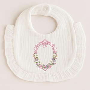 May include: White baby bib with a ruffled edge and a floral embroidery design. The embroidery features a pink oval frame with a bow at the top and a wreath of flowers in shades of purple, yellow, and white.