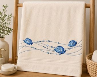 May include: A cream-colored bath towel featuring an embroidered design of three blue fish swimming among wavy lines and small circular bubbles. The towel has a textured fabric surface and a horizontal band detail near the bottom edge.