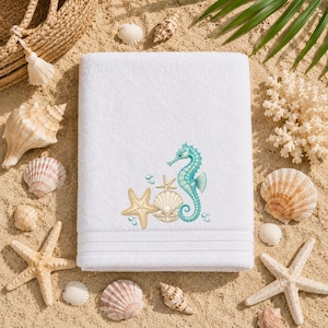 Può includere: Asciugamano da spiaggia bianco con ricamo di cavalluccio marino, stelle marine e conchiglie nei toni del blu, beige e crema. L'asciugamano è circondato da conchiglie e sabbia, evocando un tema costiero.