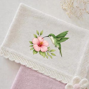 Puede incluir: Una manta de bebé de punto blanco con un borde festoneado y un diseño floral. La manta presenta una ilustración detallada de un colibrí verde que se cierne cerca de una flor de hibisco rosa con hojas verdes. La manta es suave y acogedora.