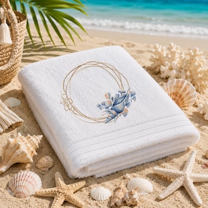 Puede incluir: Toalla de playa blanca con un diseño bordado. El diseño presenta un marco circular dorado y una concha con acentos azules y melocotón. La toalla está doblada sobre arena, rodeada de conchas marinas y estrellas de mar.