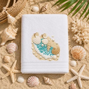 May include: A white bath towel featuring an embroidered design of tan seashells, white pearls, and a turquoise ocean wave. The towel has a soft, plush texture with a horizontal ribbed border detail near the bottom edge of the fabric.