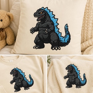 May include: A cream-colored throw pillow, a cream-colored sweatshirt, and a cream-colored fleece blanket, each featuring an embroidered graphic of a black monster with blue dorsal fins and a blue tail, standing in a bipedal stance.