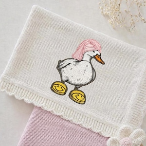 Peut inclure: Une couverture pour bébé en tricot blanc avec un bord festonné et un motif brodé fantaisiste. Le motif représente un canard de dessin animé portant un chapeau rose et des chaussures jaunes avec des visages souriants. Une deuxième couverture rose est visible en dessous.