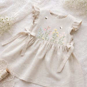 May include: A beige linen dress with embroidered flowers in pastel colors and two small bees. The dress features ruffled sleeves and tie closures at the sides. The dress is designed for a child.