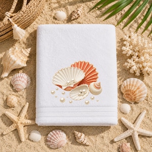 May include: A white rectangular bath towel featuring an embroidered design of two open scallop shells in cream and orange, containing several white pearls, with additional scattered pearl accents surrounding the shells on the fabric surface.