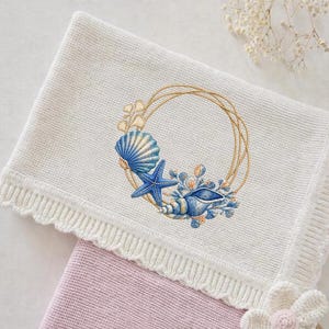 May include: Cream knitted baby blanket with an embroidered seashell design. The design features a gold geometric frame with blue seashells, a starfish, and floral accents. The blanket has a scalloped edge.