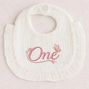 Peut inclure: Bavoir de bébé blanc avec bordure à volants et fermeture à bouton. Le bavoir porte le mot "One" brodé en rose, avec un papillon et des motifs floraux. Fabriqué en tissu doux et texturé.