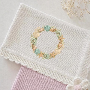 May include: A cream-colored knitted blanket with a detailed embroidered wreath of seashells, starfish, and pearls. The wreath is in shades of beige, green, and blue. The blanket has a scalloped edge and is partially covering a pink blanket.