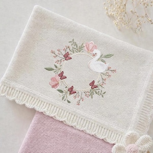 Op de afbeelding: Crèmekleurige gebreide babydeken met een bloemen- en zwanenborduurontwerp. De borduursels tonen een krans van roze bloemen, vlinders en een witte zwaan. De deken heeft een geschulpte rand.