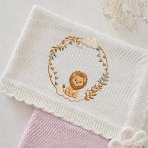 Peut inclure: Une couverture de bébé blanche tricotée avec un motif de lion brodé. Le lion est assis sur un nuage dans une couronne de feuilles et de petits drapeaux. La couverture a un bord festonné et est superposée à une couverture rose.