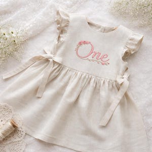 May include: A cream-colored linen dress with ruffled sleeves and tie closures. The word "One" is embroidered in pink with a floral wreath design. The dress is designed for a baby's first birthday.