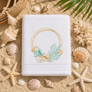 Könnte beinhalten: Weißes Strandtuch mit einem gestickten Design aus Muscheln, einem Seestern und Korallen in einem goldenen kreisförmigen Rahmen. Das Handtuch ist von Muscheln und Seesternen auf sandigem Untergrund umgeben, was ein Küstenmotiv hervorruft.