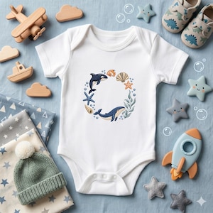 Op de afbeelding: Witte babybody met een geborduurd zeethema-ontwerp met walvissen, zeesterren en schelpen. Het ontwerp is geborduurd in blauw, oranje en beige. De afbeelding bevat ook houten speelgoed, een gebreide muts en babyschoenen.