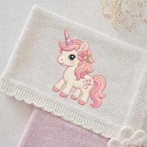 Pode incluir: Um cobertor de bebê branco de malha com um design de unicórnio rosa e lavanda. O unicórnio tem uma crina, cauda e detalhes florais rosa. O cobertor tem uma borda ondulada e é sobreposto a um cobertor rosa.