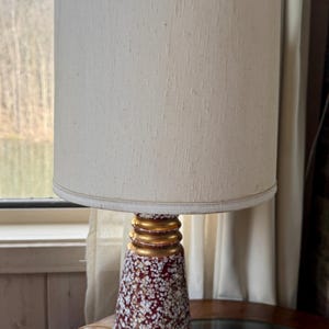 May include: A vintage table lamp with a cream-colored lampshade and a speckled red and white ceramic base. The base has gold-colored accents. The lamp is on a round table near a window.