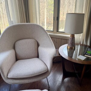 May include: A mid-century modern style living room with a light beige armchair, a round side table with a lamp, and a window with sheer curtains. The lamp has a white shade and a patterned base. A book is on the table.