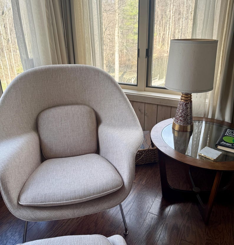 May include: A mid-century modern style living room with a light beige armchair, a round side table with a lamp, and a window with sheer curtains. The lamp has a white shade and a patterned base. A book is on the table.
