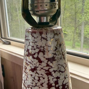 May include: A vintage lamp with a white and burgundy speckled ceramic base, accented with gold. The lamp has a silver metal socket and a black on/off switch. The lamp is positioned near a window with a view of trees.