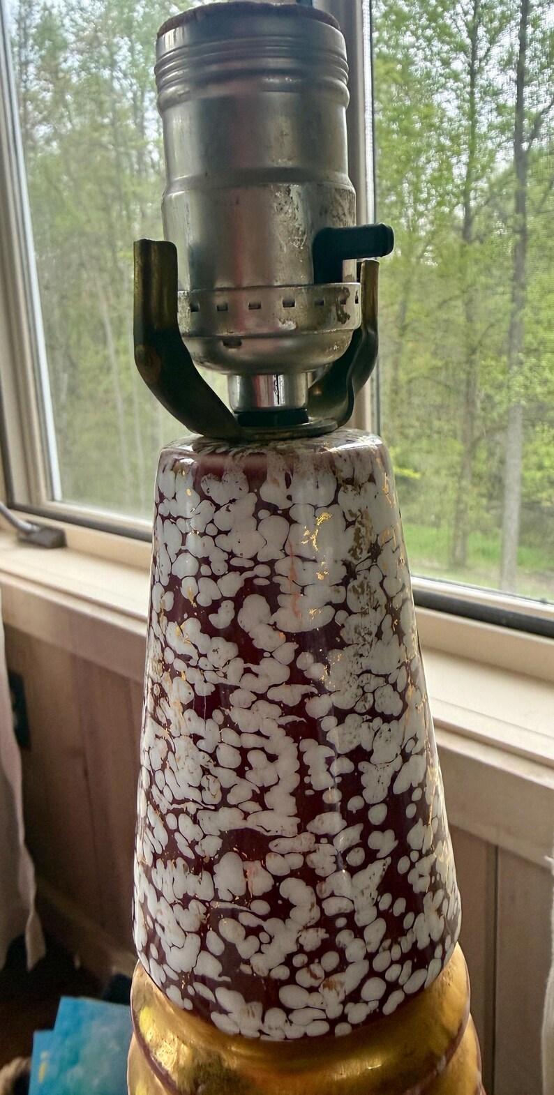 May include: A vintage lamp with a white and burgundy speckled ceramic base, accented with gold. The lamp has a silver metal socket and a black on/off switch. The lamp is positioned near a window with a view of trees.