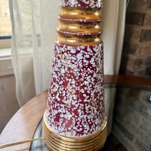 May include: A vintage table lamp with a burgundy base speckled with white and gold accents. The lamp features gold bands and a textured design. The lamp is on a wooden table.