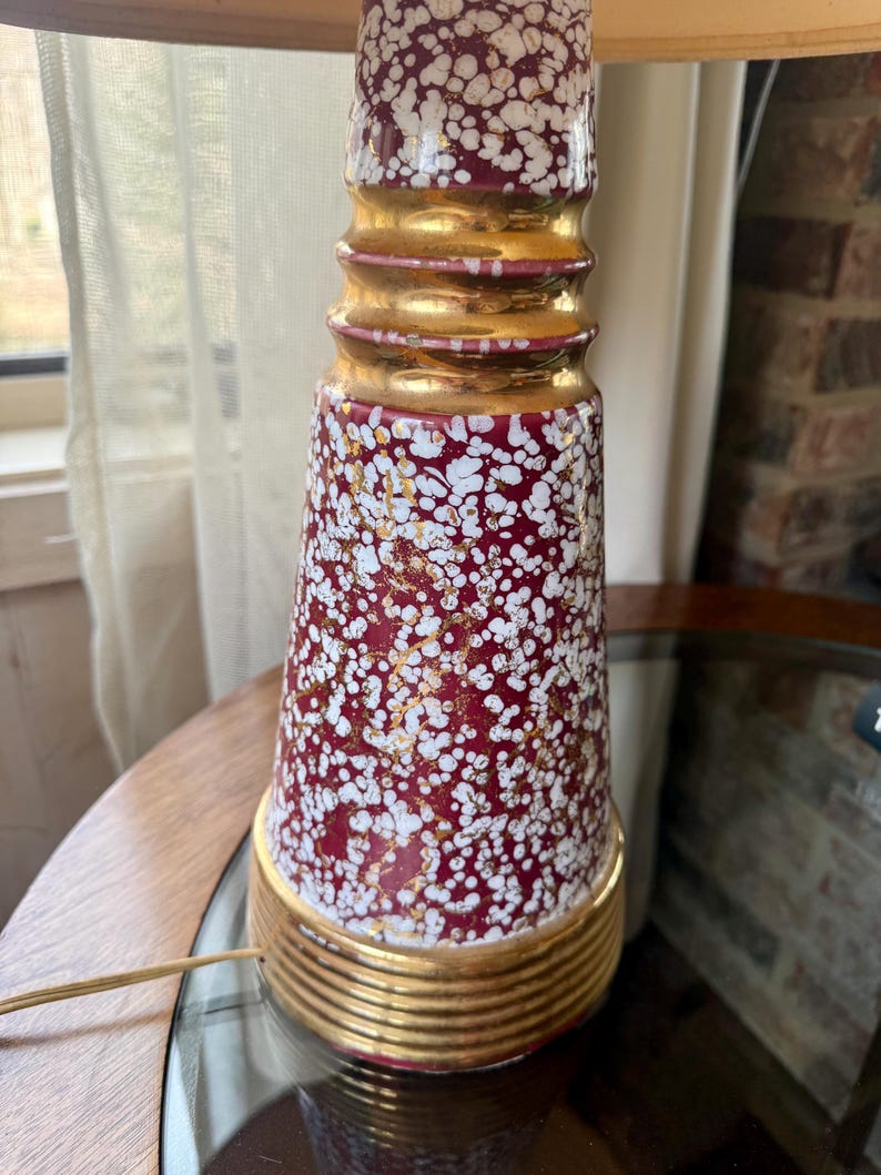 May include: A vintage table lamp with a burgundy base speckled with white and gold accents. The lamp features gold bands and a textured design. The lamp is on a wooden table.