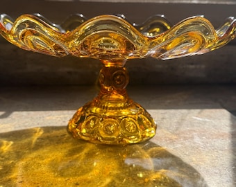 May include: A translucent amber glass pedestal bowl featuring a scalloped rim and a textured pattern of raised circles and stars on the base and stem. The bowl has a wide, shallow basin and a sturdy, circular footed stand.