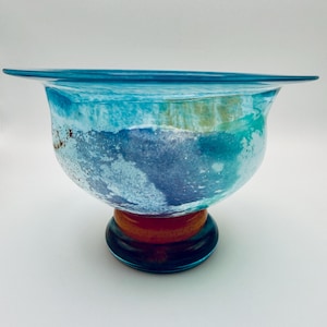 May include: A glass bowl featuring a wide, flared rim and a rounded body with a mottled pattern of blue, turquoise, and white hues. The bowl is supported by a thick, circular base that transitions from a deep orange to a dark blue glass.