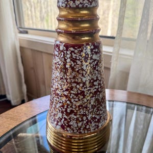 May include: A vintage table lamp with a burgundy and white speckled ceramic base, accented with gold bands. The lamp sits on a glass-topped table. The lamp's shade is a light beige color.