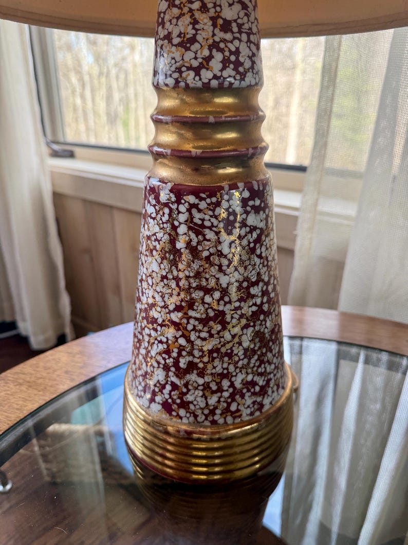 May include: A vintage table lamp with a burgundy and white speckled ceramic base, accented with gold bands. The lamp sits on a glass-topped table. The lamp's shade is a light beige color.
