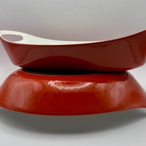 May include: Two oval-shaped baking dishes with a glossy orange exterior and a smooth white interior. Each dish features integrated handles on both ends, with one dish stacked directly on top of the other, showcasing the vibrant orange finish.