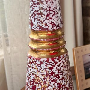 May include: A decorative table lamp with a red base covered in white speckles and gold accents. The lamp has a gold base and a cylindrical shape, with a standard lamp socket at the top. The lamp is a vintage or retro style.