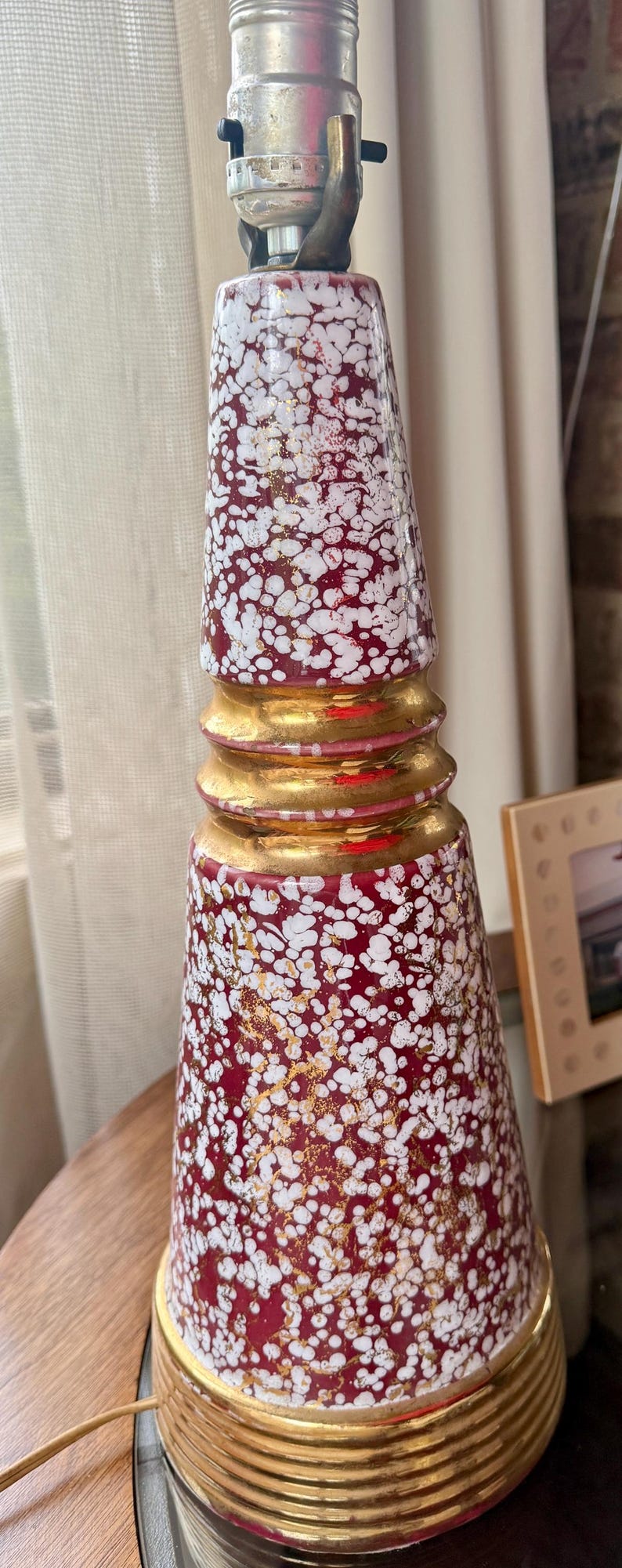 May include: A decorative table lamp with a red base covered in white speckles and gold accents. The lamp has a gold base and a cylindrical shape, with a standard lamp socket at the top. The lamp is a vintage or retro style.