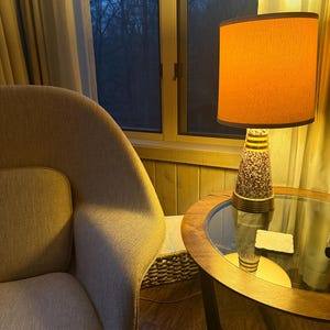 May include: A mid-century modern lamp with an orange shade and a patterned base sits on a round glass-topped table. A beige armchair is in the foreground, and a window with a view of trees is in the background.