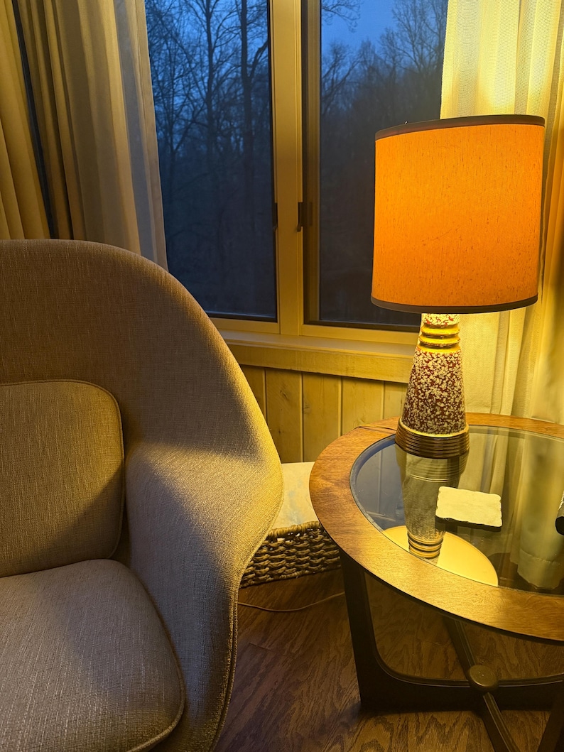 May include: A mid-century modern lamp with an orange shade and a patterned base sits on a round glass-topped table. A beige armchair is in the foreground, and a window with a view of trees is in the background.