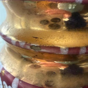 May include: Close-up of a decorative glass object with a gold metallic finish. The object has a bulbous shape with red and white accents. The design includes a series of circular patterns and a textured surface.