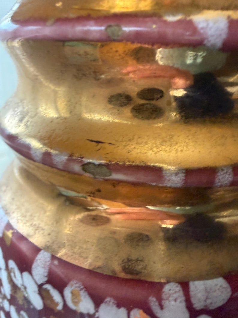 May include: Close-up of a decorative glass object with a gold metallic finish. The object has a bulbous shape with red and white accents. The design includes a series of circular patterns and a textured surface.