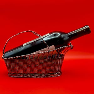 May include: A dark brown metal wire basket with a woven pattern and a curved handle, holding a dark glass bottle positioned at an angle. The bottle features a white rectangular label with visible text, resting securely within the wire structure.