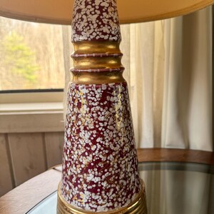 May include: A vintage table lamp with a burgundy and white speckled ceramic base, accented with gold bands. The lamp has a cream-colored lampshade. The lamp is on a mirrored surface.
