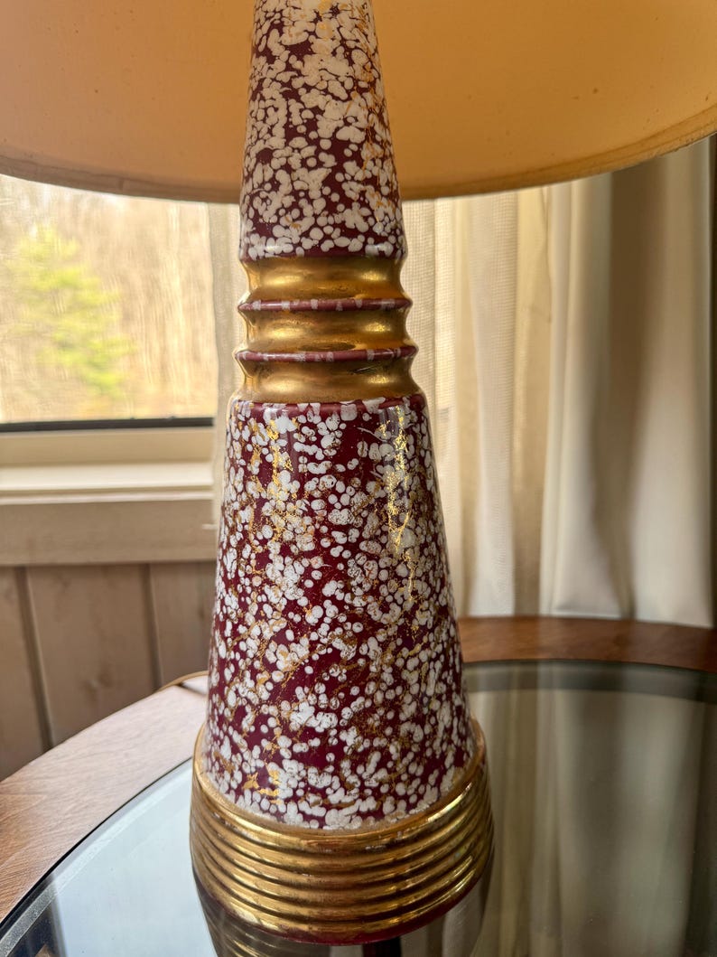 May include: A vintage table lamp with a burgundy and white speckled ceramic base, accented with gold bands. The lamp has a cream-colored lampshade. The lamp is on a mirrored surface.