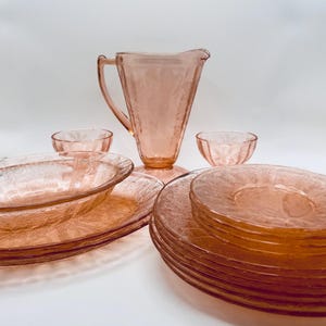 May include: A collection of pink glassware, including a pitcher, bowls, and plates. The set features a textured pattern and is arranged on a white surface. The glassware is likely for serving or display.