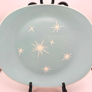 May include: A round ceramic plate with a smooth light blue finish featuring a pattern of white starburst designs of varying sizes scattered across the center. The plate has a clean, simple edge and is displayed on a black metal stand.