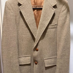 May include: A tan and brown tweed blazer with a classic two-button closure and notched lapels. The jacket has two front pockets and a chest pocket. The interior lining is a solid brown color. The blazer is hanging on a wooden hanger.