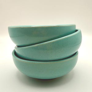 May include: A stack of three turquoise ceramic bowls. The bowls have a speckled texture and a slightly rounded shape. The rims of the bowls have a lighter, tan-colored edge. The bowls are stacked on top of each other.