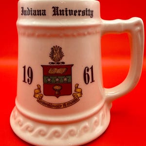 May include: A white ceramic beer stein featuring the text Indiana University at the top, the numbers 19 and 61 on the sides, and a central crest with a book, stars, and wheat. The base and rim have a raised wave pattern, and a handle is on the right.