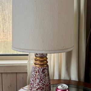 May include: A vintage table lamp with a cream-colored lampshade and a decorative base. The base is a deep red color with white and gold accents. The lamp is on a round glass table. A can of Waterloo Cherry Limeade is next to the lamp.